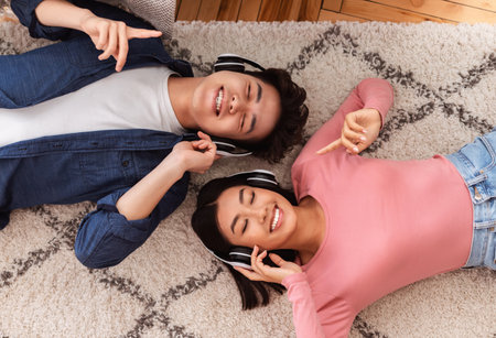 Cheerful Asian Boyfriend And Girlfriend Wearing Headphones Listening To Music Lying On Floor With Eyes Closed Relaxing At Home. Couple Enjoying Favorite Romantic Songs On Weekend. View From Aboveの写真素材