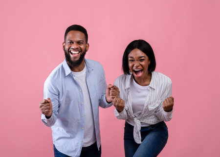 Lucky winners. Emotional black couple celebrating success, making YES gesture, yelling in excitement, pink studio background. Young African American guy and his girlfriend rejoicing togetherの写真素材