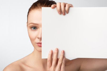 Red-Haired Young Woman Holding White Poster Covering Half Of Face Posing Looking At Camera Standing Over White Studio Background. Female Beauty Portrait With Copy Spaceの写真素材