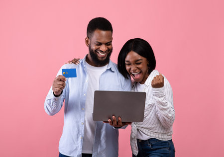 Huge sales in webstore. Excited black woman and her boyfriend shopping online with credit card and laptop on pink studio background. African American millennials purchasing goods on internetの写真素材