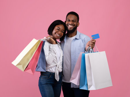 Positive African American couple with credit card and shopping bags smiling and posing on pink studio background. Millennial guy and his girlfriend purchasing Valentines gifts remotelyの写真素材