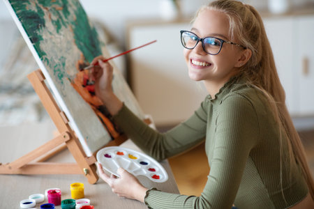 Inspired blonde teen girl with brush and palette painting, smiling at camera home interior. Creative schooler enjoying her new hobby, painting still life, side view. Teenagers lifestyle conceptの写真素材