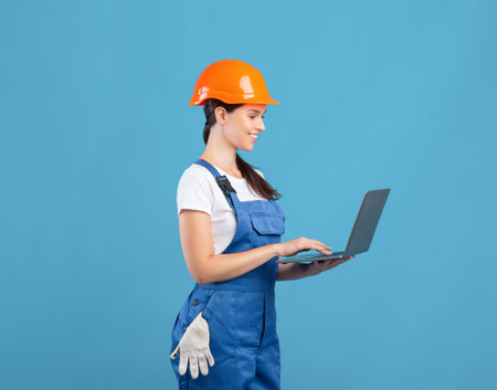 Beautiful Young Female Maintenance Worker Using Laptop, Ordering Construction Materials Online. Lady In Hardhat Making Repair Tools Shopping In Internet, Standing On Blue Background, Copy Spaceの写真素材