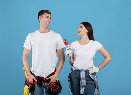 Young couple with drill and safety glasses ready to start repair, millennial spouses standing with construction tools over blue studio background and smiling to each other, copy spaceの写真素材