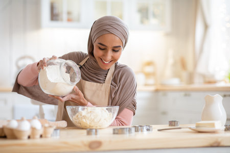 Happy muslim woman in hijab baking in kitchen, preparing dough for cookies, adding flour to bowl, enjoying making homemade pastry, beautiful islamic lady in headscarf cooking at home, free spaceの写真素材
