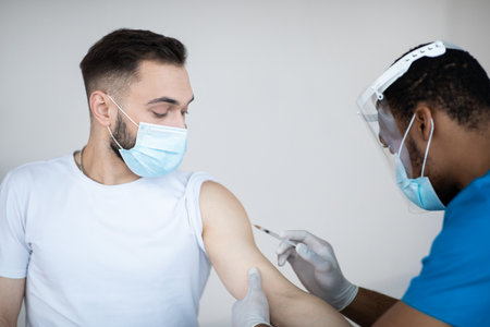 Young Caucasian man getting injection of coronavirus vaccine at hospital. African American medical worker immunizing male patient against covid-19 at clinic. Healthcare and global infection preventionの写真素材