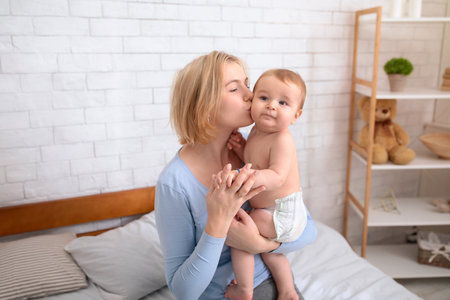 Young blonde mother kissing her adorable little kid son or daughter, bedroom interior, free space. Loving mom and cute baby boy or girl in diaper, mummy waking up her sweet babyの写真素材