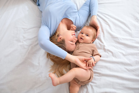 Adorable baby boy and loving blonde mother laying on bed head to head, top view. Joyful young woman playing with her cute little kid on bed, laying and bonding together at homeの写真素材