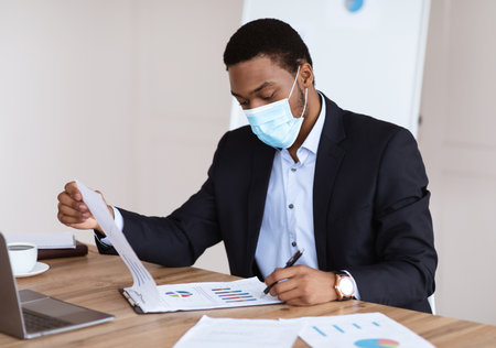 Young black manager in face mask reading marketing report and signing, sitting at his workplace in front of laptop in office, african businessman doing paperwork during covid-19 pandemic, copy spaceの写真素材