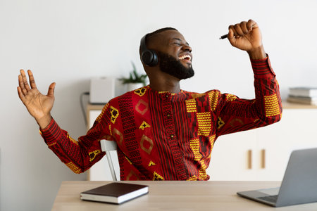 Break In Work. Cheerful African Guy In Traditional Shirt Singing At Workplace, Using Pen As Microphone And Listening Music In Wireless Headphones, Joyful Freelancer Guy Relaxing At Desk In Home Officeの写真素材