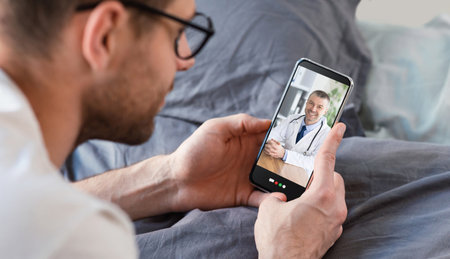 Young man having online consultation with doctor on smartphone, using telemedicine app on bed, panorama. Male patient communicating to his physician on mobile device from homeの写真素材
