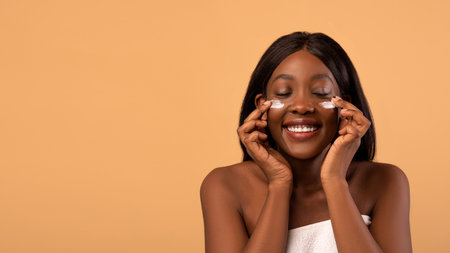 Closeup of beautiful young african american woman with closed eyes smiling while applying hydrogel eye patches after shower, beige studio background, panorama with copy spaceの写真素材