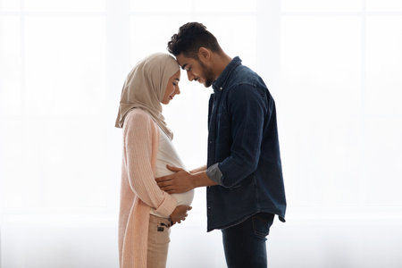 Young religious muslim couple expecting baby, standing together near window at home, caring millennial arab husband tenderly touching belly of his pregnant wife in hijab, side view with copy spaceの写真素材