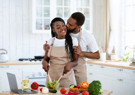 Romantic Black Couple Kissing While Cooking Healthy Food Together In Kitchen. Affectionate African American Spouses Preparring Vegetarian Lunch On Table With Laptop, Enjoying Time Together, Free Spaceの写真素材