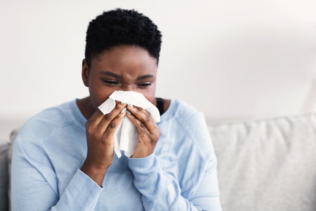 Sick african american woman sneezing, holding tissue paper napkin, sitting on the couch at home. Young ill black lady got cold or flu, having fever and suffering from runny nose, copy spaceの写真素材