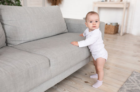 Adorable Little Baby Looking At Camera Standing Near Couch At Home. Cute Toddler Making First Steps In Living Room Alone. Childhood Concept. Blank Space For Textの写真素材