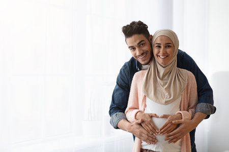 Portrait of happy pregnant religious muslim couple posing near window at home. Loving arab husband hugging his expecting wife in hijab from behind, making heart shaped sign on belly, copy spaceの写真素材