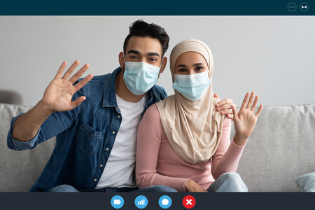 Muslim couple in protective face masks waving at camera, having video conference with friends or family while pandemic, laptop or digital tablet webcam screen. Technologies in communicationの写真素材