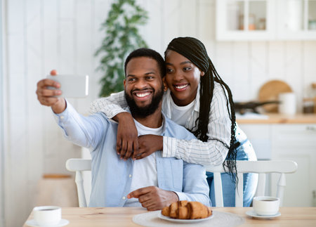 Romantic African American Couple Taking Selfie On Smartphone While Having Breakfast In Kitchen Together. Happy Black Spouses Embracing And Posing At Camera, Enjoying Morning At Home, Free Spaceの写真素材