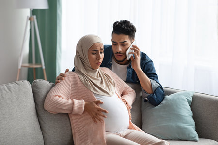 Worry Muslim Husband Calling Emergency While His Pregnant Wife Having Prenatal Contractions At Home. Islamic Expecting Mother In Hijab Suffering Painful Spasms And Spontaneous Labour, Copy Spaceの写真素材