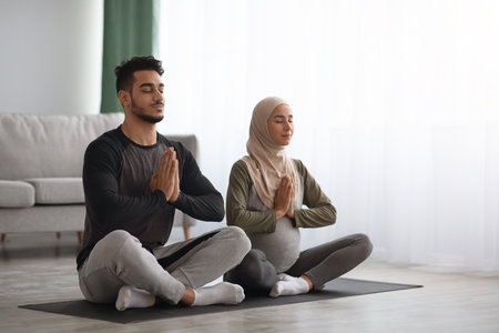 Pregnant Muslim Woman In Hijab Meditating Together With Her Husband At Home. Arab Couple Practicing Prenatal Yoga In Living Room, Sitting With Clasped Hands, Enjoying Healthy Lifestyle, Copy Spaceの写真素材