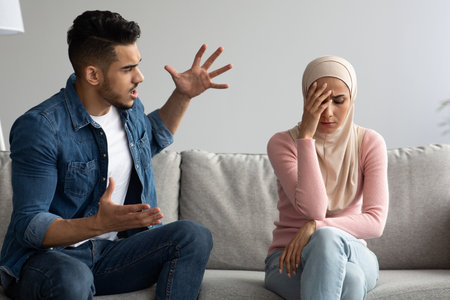 Relationships problems and crisis concept. Angry arab man and woman in hijab sitting on couch at home, having quarrel, copy space. Furious middle eastern guy yelling at his upset girlfriendの写真素材