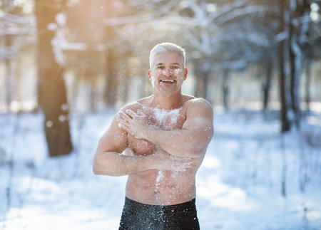 Athletic mature guy tempering his body with snow, developing resistance to cold at white winter forest. Strong senior man with bare torso doing acclimation procedures. Seasonal activities conceptの写真素材