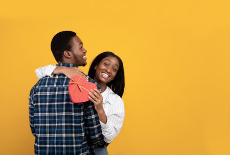 Happiness together, anniversary, Valentines day and love. Smiling young african american wife hugging husband and holding box in heart shaped, isolated on yellow background, studio shot, free spaceの写真素材