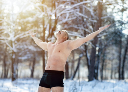Happy mature man holding his arms out, standing in underwear in winter frost. Healthy senior guy doing cold resistance training, feeling freedom at snowy forest. Seasonal activitiesの写真素材