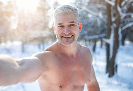 Smiling mature guy with naked torso taking selfie at snowy winter park. Happy senior man taking photo of himself while doing cold exposure training outdoors. Seasonal pastimesの写真素材