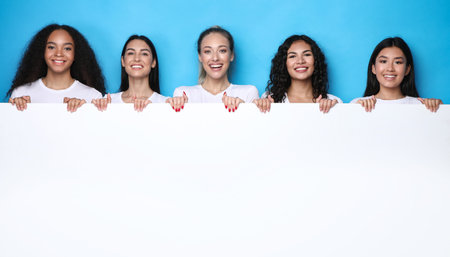 Group Of Five Diverse Women Holding Big Paper Poster For Your Text Standing Over Blue Studio Background. Multicultural Females Showing Blank White Board For Advertisementの写真素材