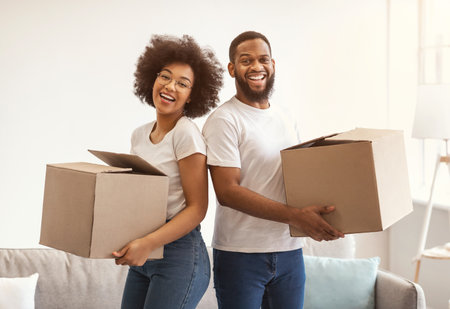 Joyful Black Couple Moving House Posing Holding Packed Boxes Preparing For Relocation Standing Indoors, Smiling To Camera. Real Estate Purchase, Mortgage And Ownershipの写真素材