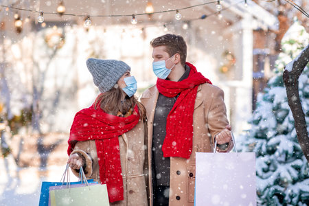 Family enjoying seasonal discounts and huge sales, empty space. Young couple in protective face masks holding colorful shopping bags, man and woman purchasing together on winter snowy day