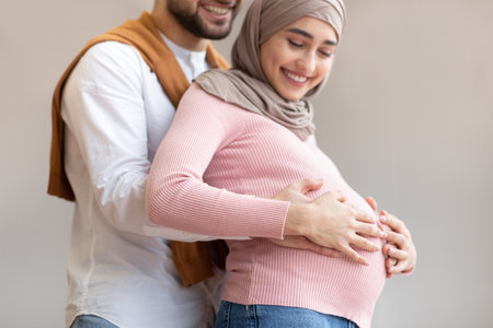 Pregnancy. Happy Muslim Couple Waiting For Baby, Arab Husband Hugging Pregnant Wifes Belly Posing On Gray Studio Background. Family Happiness And New Life. Cute Parents-To-Be Portrait. Croppedの写真素材
