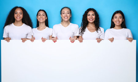 Five Multicultural Women Holding Empty White Board Advertising Your Text Smiling To Camera Standing On Blue Background In Studio. Females Group Showing Big Empty Poster For Advertisement. Mockupの写真素材