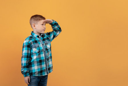 Cute ginger kid in flannel holding palm next to forehead, looking at copy space, panorama, yellow studio background. Curious red-haired boy watching exciting advertisement for kidsの写真素材