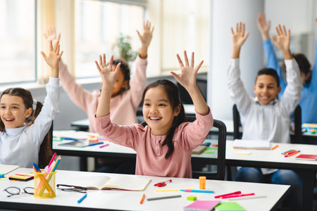 Elementary School and People Concept. Diverse excited group of emotional happy junior school kids sitting at desks in classroom and raising hands, having fun, studying with pleasureの写真素材