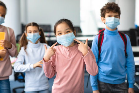 Protection Concept. Portrait of asian girl wearing medical surgical face mask to prevent infection, illness, flu or covid-19, pointing at her covered mouth standing in classroom at school. Pandemicの写真素材