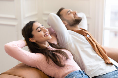 Happy Young Arab Couple Relaxing Sitting On Sofa Holding Hands Behind Head Enjoying Lazy Weekend Together At Home. Millennials Spouses Enjoying Resting On Couch With Eyes Closed. Selecitve Focusの写真素材