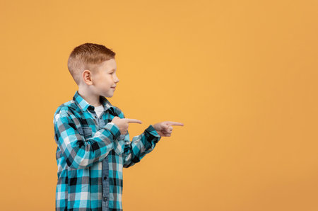 Curious ginger boy in casual outfit pointing with both hands at empty space for text or advertising over yellow studio background. Smiling kid showing exciting advertisement, aiming at free spaceの写真素材