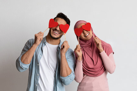 Valentines Fun. Cheerful Muslim Man And Woman Covering Eyes With Red Paper Hearts, Playful Islamic Couple In Love Posing Together Over Grey Background In Studio, Girlfriend Wearing Hijab, Copy Spaceの写真素材