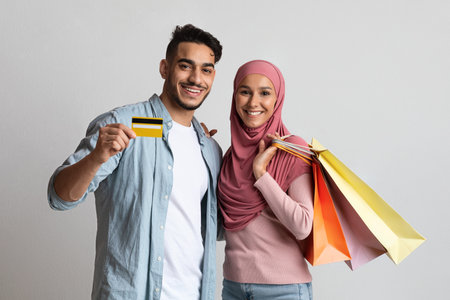 Reliable Bank. Cheerful muslim couple posing with credit card and shopping bags, young arab man and islamic woman in hijab enjoying easy payments while making purchases, standing on grey backgroundの写真素材