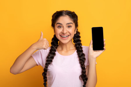 I Recommend. Portrait of excited young indian woman holding smartphone with black blank screen in hand, showing device and thumbs up sign gesture. Gadget with empty free space for mock upの写真素材