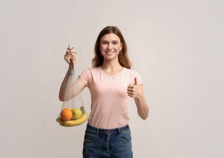Beautiful Smiling Woman Holding Washable Mesh Net String Bag With Organic Fruits And Showing Thumb...