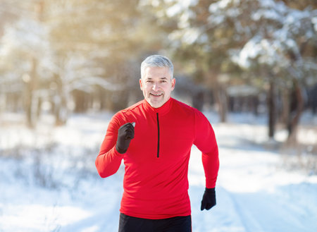 Cross country running in winter. Happy fit senior man jogging in snowy forest on sunny morning. Sporty mature guy going for run in frosty weather, getting ready for race or marathonの写真素材
