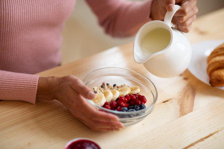 Healthy Breakfast. Unrecognizable Woman Eating Muesli With Fruits In The Morning, Sitting At Table Holding White Jug And Pouring Milk To Bowl, Enjoying Delicious Meal, Cropped Image, Closeupの写真素材