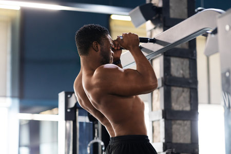 Muscular black guy athlete pulling up on horizontal bar at gym, free space. Shirtless african...