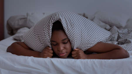 Neighbors interfere to sleep, noise, nightmares, problems and bad thoughts. Upset young african american lady with closed eyes covers her head with pillow on bed in bedroom interior, dark, panoramaの写真素材