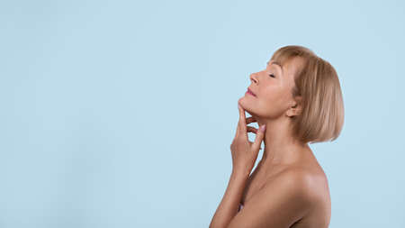 Graceful aging concept. Beautiful senior lady touching skin on her neck over blue studio background....