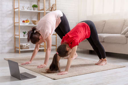 Home sports during covid. Young mother and her daughter exercising at home to online video on laptop, standing in downward facing dog pose. Parent and kid doing domestic yogaの写真素材